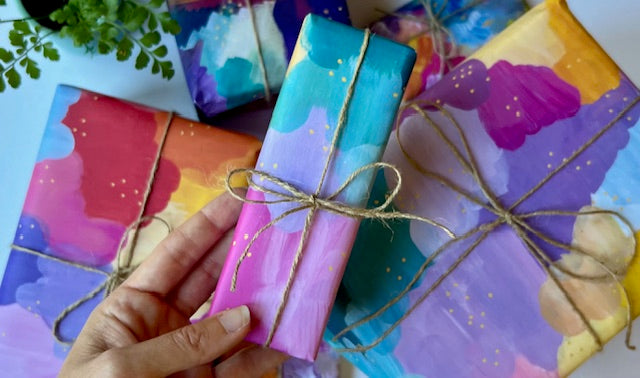 A hand holds a wrapped gift box with a colorful painted design in shades of pink, purple, and blue, tied with twine. Other similarly wrapped gift boxes are visible in the background on a light surface. Artist made paintings in wrapping paper colored with aqua, pink and yellow.