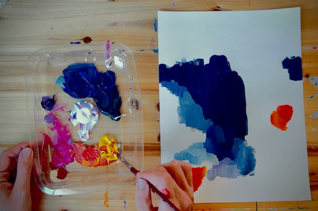 A top-down view of a hand holding a paintbrush next to a clear plastic palette filled with colorful paint, with a white sheet of paper showing abstract painted swatches on a wooden table.